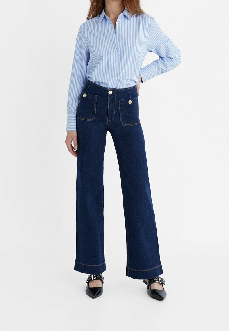 Femme portant une chemise rayée bleu clair boutonnée rentrée dans un jean large taille haute bleu foncé avec des poches plaquées devant et des chaussures pointues noires.