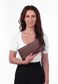 Hnědá kožená kabelka na peníze s překlopným designem a embosovaným logem, držena v ruce na bílém pozadí.