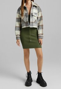 Krótka kurtka w kratę w odcieniach szarości, bieli i brązu, wykonana z mieszanki wełny, z dwiema kieszeniami na piersi; zielona sukienka z prążkowanego materiału; czarne, sznurowane buty na platformie.