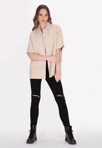 Camicia beige a maniche corte in materiale scamosciato sintetico, con colletto e bottoni a pressione, abbinata a jeans skinny neri strappati e stivali neri con lacci.