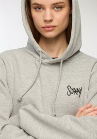 Felpa grigia realizzata in materiale morbido, con cappuccio regolabile e logo "SOARY" stampato in nero sul davanti. Design semplice.
