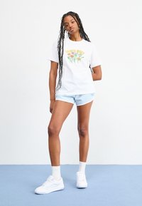 T-shirt en coton blanc avec un design floral et le texte "SANTA CRUZ", associé à un short bleu clair. Le modèle porte des baskets blanches et des chaussettes.