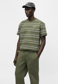 Άνδρας που φοράει πράσινη ριγέ μπλούζα, χακί παντελόνι με τσέπες και σκουρόγκρι καπέλο, στέκεται με το ένα χέρι στην τσέπη.