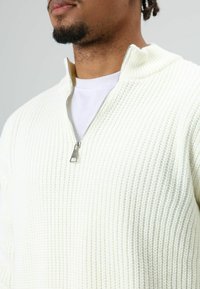 Man wearing white textured knit sweater partially unzipped over white crew neck shirt, with close-up on neck and lower face.