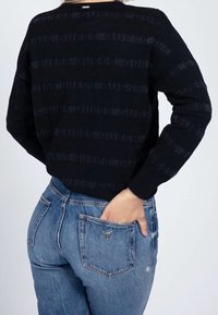 Černý strukturovaný svetr s vodorovnými pruhy, zkombinovaný s světle modrými džínami, které mají trojúhelníkové logo na zadní kapse.