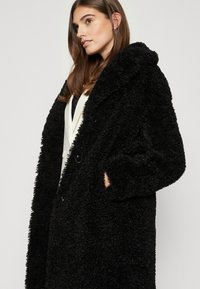 Cappotto nero e morbido con finitura testurizzata, caratterizzato da un colletto a scialle, due bottoni e tasche laterali. Indossato da un modello su uno sfondo neutro.