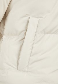 Beige pufferjakke med et glat yderstof, syet mønster og en sidelomme med klap. Blød, isoleret tekstur.