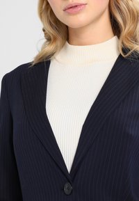 Marineblauw blazer met krijtstreep en een gekerfde kraag, gecombineerd met een geribbelde crèmecoltrui. Heeft een enkele knoopsluiting en een gladde stoftextuur.