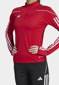 Rotes sportliches Langarmoberteil mit halb durchgehendem Reißverschlusskragen, weißen Akzenten und drei weißen Streifen an jedem Arm; strukturierte Schultern; feuchtigkeitsableitendes Material.