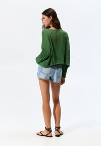Grüner gestrickter Pullover mit lockerem Schnitt und strukturiertem Design, kombiniert mit hellblauen, verwitterten Jeansshorts und braunen, Riemchensandalen.