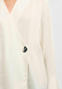 Cremefarbene Wickelbluse mit langen Ärmeln, ausgestattet mit einem glänzenden schwarzen Knopfakzent und einer weichen, leichten Stofftextur.