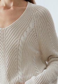 Beige stickad tröja med djup V-ringning, med ett kabelstickat mönster och texturerat stygn detalj längs sömmarna. Mjuk, lättviktsmaterial.