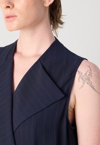 Prenda sin mangas de color azul marino con un cuello afilado, que presenta sutiles rayas finas y una textura suave. Se puede ver un tatuaje en el brazo.