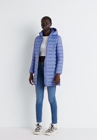 Pěnový kabát ve světle fialové barvě, se sklápěcím límcem, zipovým zapínáním vpředu a vertikálním prošíváním. Nosený přes bílý crop top a modré džíny.