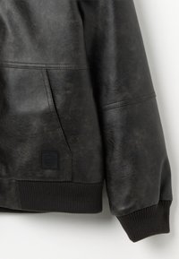Chaqueta de cuero negra con una superficie texturizada, puños acanalados y un bolsillo lateral con un parche de logo cuadrado.