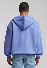 Hanorac oversized violet cu glugă reglabilă, mâneci și tiv cu nervuri, realizat dintr-o țesătură moale de bumbac. Purtați cu blugi din denim albastru deschis.