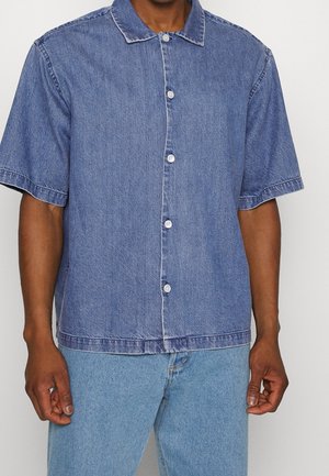 Mann trägt ein kurzärmeliges blaues Jeanshemd mit Kragen und hellblaue Jeans vor schlichtem Hintergrund.