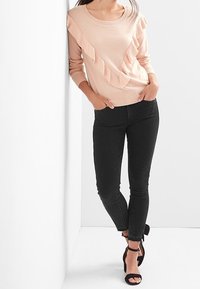 Helles rosa Sweatshirt mit Rüschendetails, kombiniert mit schwarzen Skinny-Jeans und schwarzen High Heels mit Knöchelriemen; Modell lehnt sich an eine weiße Wand.