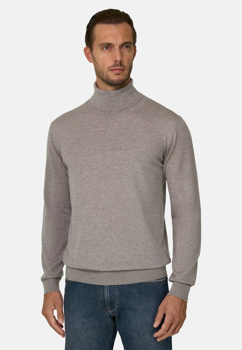 Homme portant un pull à col roulé gris clair et un jean bleu, debout avec une expression neutre sur un fond uni.