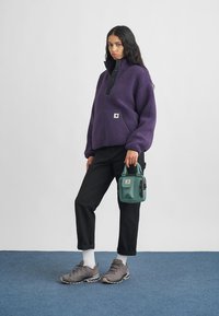 Paarse fleece trui met zwarte kraag, voorzak, zwarte broek, grijze wandelschoenen en een turquoise schoudertas met een logo. Witte sokken zichtbaar.