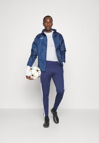 Navy windbreaker with white accents, paired with navy track pants featuring side stripes. Holding a multicolored soccer ball, wearing black cleats.