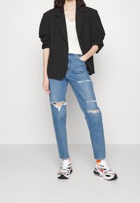 Blazer oversize à fines rayures noires, top blanc à col en V, jean bleu clair déchiré, et baskets multicolores avec des accents noirs et orange.