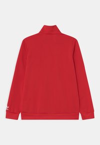 Červená bunda s vysokým límcem, dlouhými rukávy a elastickými manžetami. Hladká tkanina s logem na levém rukávu.