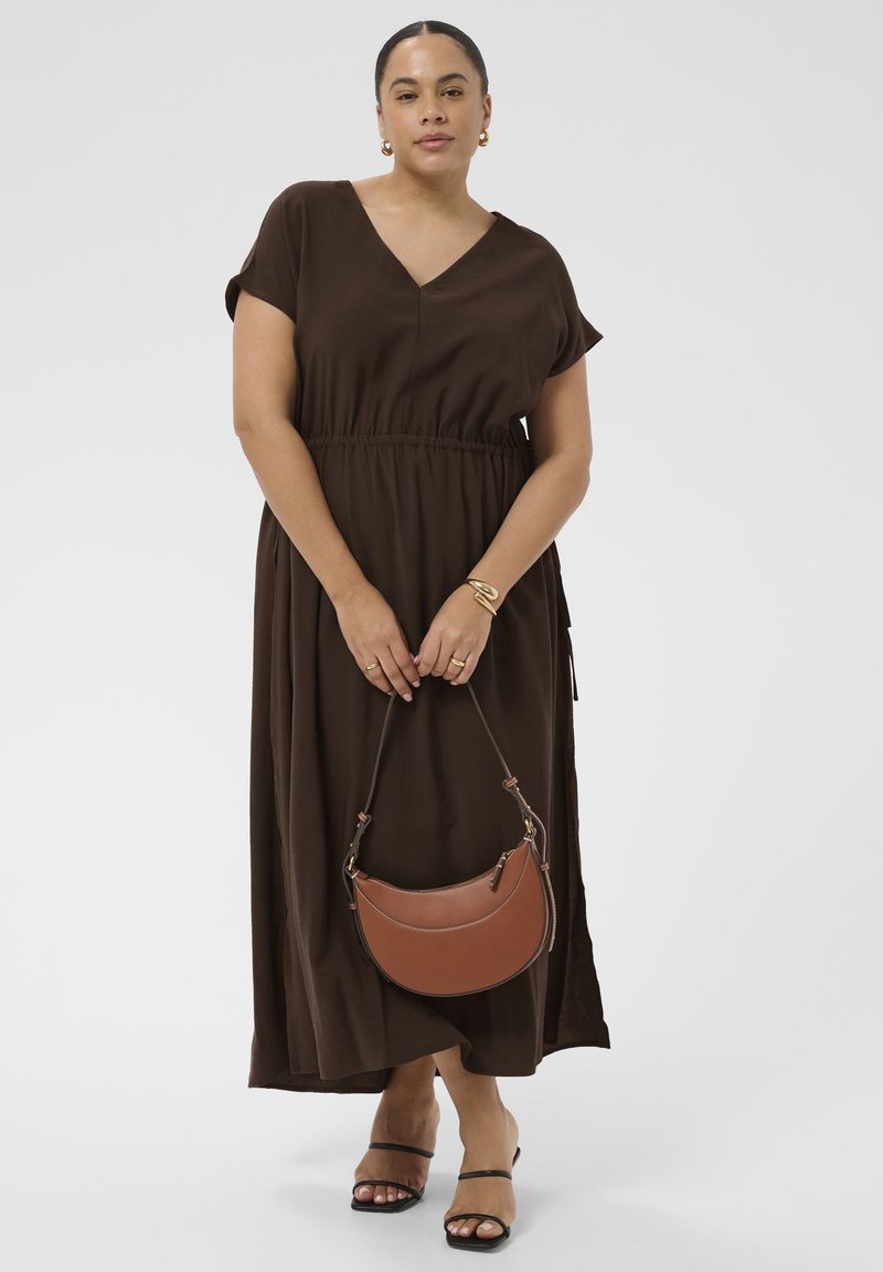 Femme en robe midi marron décolleté en V tenant un sac à main marron en forme de croissant, portant des bijoux dorés et des talons noirs à brides, debout sur fond blanc.