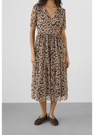 Vrouw draagt een zwarte midi-jurk met beige bloemenprint, korte mouwen, V-hals en bruine ballerina's op een grijze vloer met een witte achtergrond.