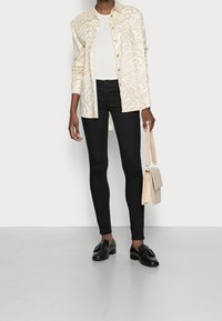 Camisa blanca con patrón zigzag beige, jeans ajustados negros, mocasines negros y un bolso beige con un diseño estructurado. Textura suave, apariencia casual.