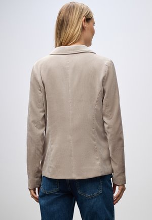 Chaqueta de pana beige con un diseño estructurado, cuello con muesca y mangas largas, llevada con jeans azules, vista desde atrás.