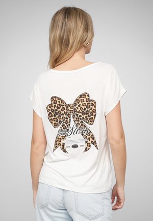 Kvinde med blondt hår iført en hvid t-shirt med en stor leopardmønstret sløjfe og sætningen "Ejer din historie" på ryggen.