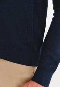 Granatowy dzianinowy sweter z prążkowanymi mankietami i zakończeniem, z boczną kieszenią i gładką fakturą. Umieszczony na beżowym tle.