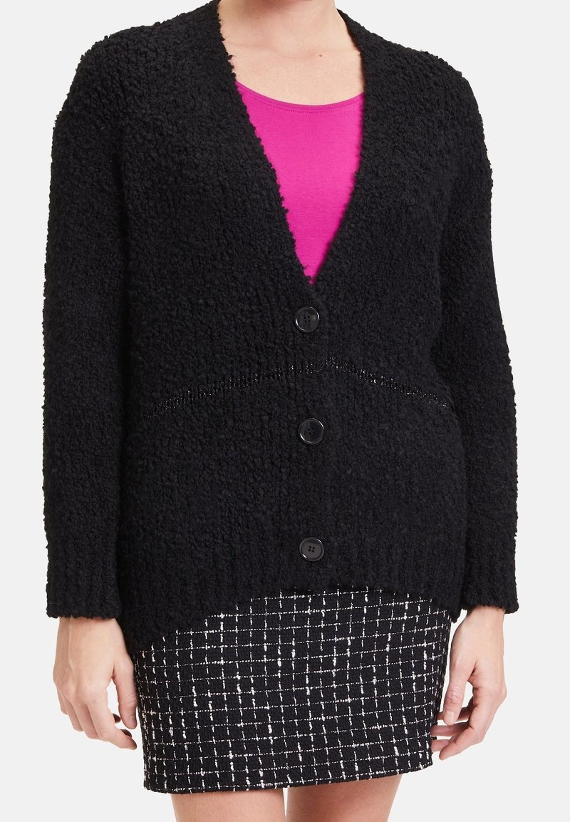 Femme portant un cardigan noir texturé boutonné sur un haut rose et une jupe à motif quadrillé noir et blanc, debout les bras détendus.