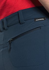 Marineblaue Hose mit strukturiertem Finish, ausgestattet mit einer Reißverschlusstasche und einem Logosticker. Der Bund hat Gürtelschlaufen und ein schlankes Design.