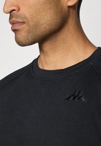 Sort sweatshirt med rund halsudskæring og tekstureret stof. Har et broderet sort logo på brystet. Glat overflade og tætsiddende design.