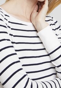 Maglia bianca a maniche lunghe con righe orizzontali blu navy. Presenta un collo a pescatore e polsini a costine, mostrando una morbida texture di cotone.