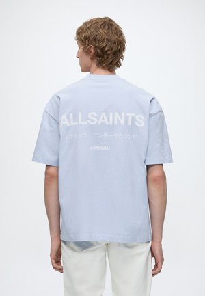 Jeune personne aux cheveux bouclés portant un t-shirt ALLSAINTS bleu clair avec des caractères japonais et un pantalon blanc, vue de dos sur fond blanc.