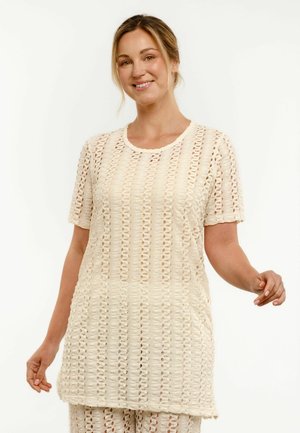 Uśmiechnięta kobieta w kremowym, krótkim rękawie, ażurowym swetrze i pasujących spodniach, stojąca na białym tle.