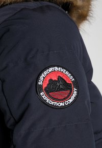 Doudoune bleu marine avec un patch circulaire noir et rouge cousu, représentant un design de montagne et le texte "Superdry Everest Expedition Company".