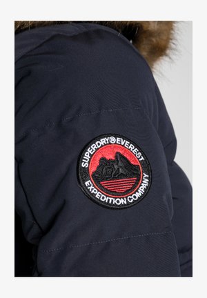 Marineblaue Steppjacke mit einem gestickten schwarz-roten runden Aufnäher, der ein Bergdesign und den Text "Superdry Everest Expedition Company" zeigt.