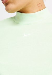 Person som bär en ljusgrön Nike-turtleneck med en liten röd hjärt-tatuering på nacken mot en vit bakgrund.