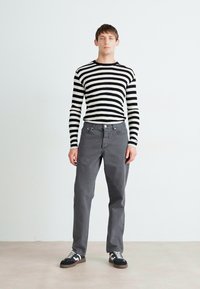 Chemise à rayures noires et blanches à manches longues portée avec un pantalon gris et des baskets noires. Fond uni aux tons neutres.