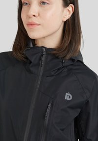Veste noire zippée avec capuche, dotée d'un tissu texturé, d'une poche zippée sur le côté et d'un logo sur la poitrine.