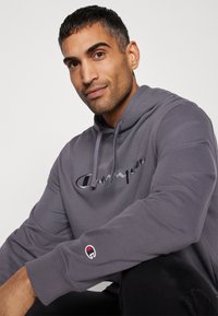 Hoodie gris avec un logo script "Champion" brodé, cordon de serrage et patch logo sur le poignet. Texture de tissu lisse.