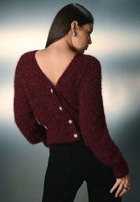 Bordeauxrød, blød sweater med dyb V-ryg, lange ærmer og dekorative knapper langs siden. Har en kort pasform og struktureret strik.