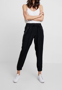 Kobieta w czarnych dresowych spodniach z troczkami i bocznymi ćwiekami, w białym topie na ramiączkach i białych sneakersach, stoi z założonymi rękami na neutralnym tle.