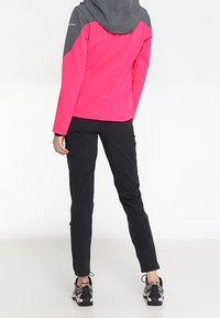 Giacca softshell rosa con dettagli grigi, cappuccio e cuciture visibili, abbinata a pantaloni aderenti neri e scarpe sportive grigie e nere.