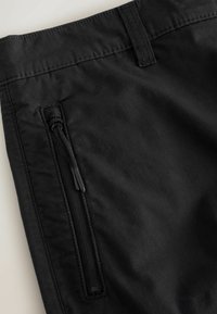 Gros plan sur un pantalon en tissu noir montrant une poche latérale zippée et une passants de ceinture avec des détails de coutures visibles.
