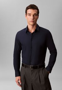 Mann mit kurzem dunklem Haar, der ein marineblaues, geknöpftes Hemd trägt, das in grau plissierte Hosen mit schwarzem Gürtel gesteckt ist, vor neutralem Hintergrund stehend.